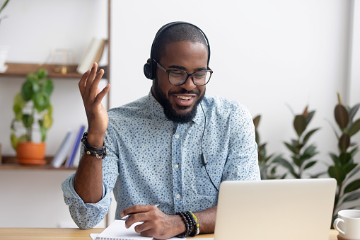 Smiling African American employee in headphones using laptop, looking at screen, making video call or watching webinar, writing notes, distance learning language concept, call center operator working