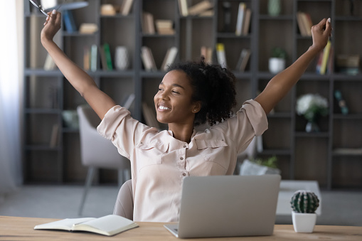 Smiling african american biracial business woman manager worker raising up arms, stretching back, relaxing after finishing hardworking day, meeting deadline, dreaming visualizing in modern home office