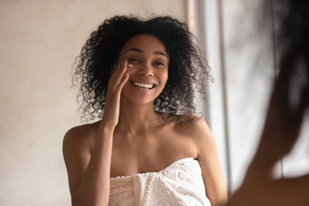 Smiling young african American woman in towel look in mirror in home...