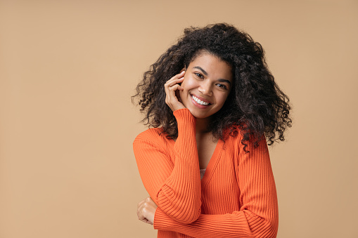 Portrait of beautiful smiling African American woman with curly hairstyle looking at camera isolated on background. Young confident fashion model wearing stylish orange sweater posing for pictures