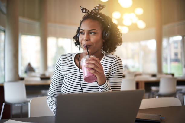Smiling African university student studying and drinking a smoothie Smiling young African college student sitting on campus drinking a smoothie and listening to music on headphones while working online with a laptop drinking smoothie stock pictures, royalty-free photos & images