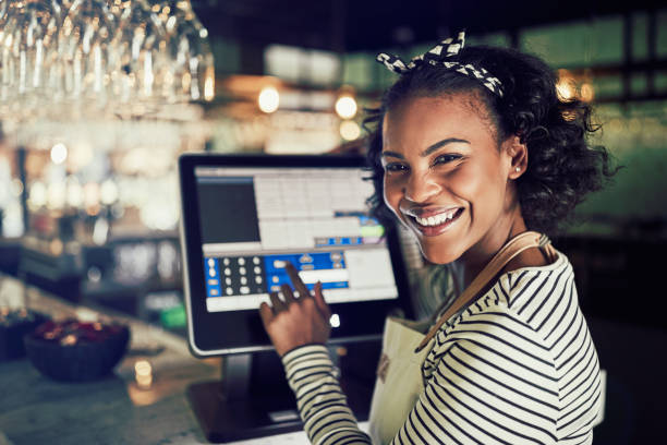 Smiling African waitress using a restaurant point of sale terminal Smiling young African waitress wearing an apron using a touchscreen point of sale terminal while working in a trendy restaurant restaurant employee pos system stock pictures, royalty-free photos & images