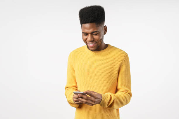 sorrindo jovem africano segurando telefone olhando para smartphone isolado no fundo de estúdio branco - afro americano - fotografias e filmes do acervo