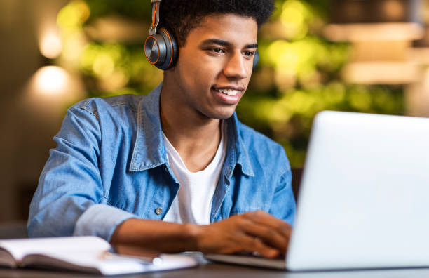 Smiling afro guy in headphones looking at laptop Smiling african american teen guy in headphones looking at laptop, studying foreign language through video conference application, cafe interior online education stock pictures, royalty-free photos & images