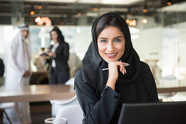 Smiling Arab businesswoman holding pen in office A photo of confident and smiling Emirati Arab businesswoman holding pen. Portrait of middle eastern professional in traditional clothing sitting with hand on chin. She is wearing abaya andworking in brightly lit modern office. middle eastern woman stock pictures, royalty-free photos & images