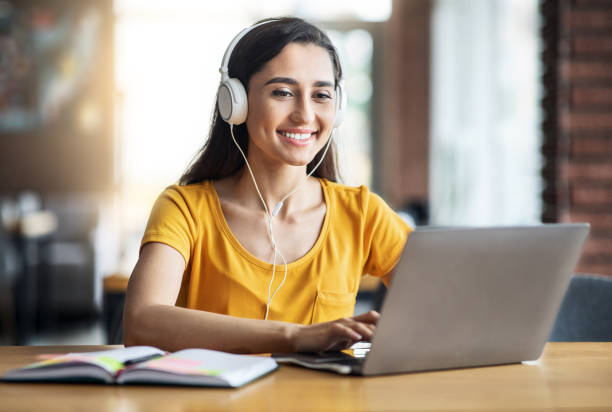 chica árabe sonriente con auriculares estudiando en línea, usando portátil - students fotografías e imágenes de stock
