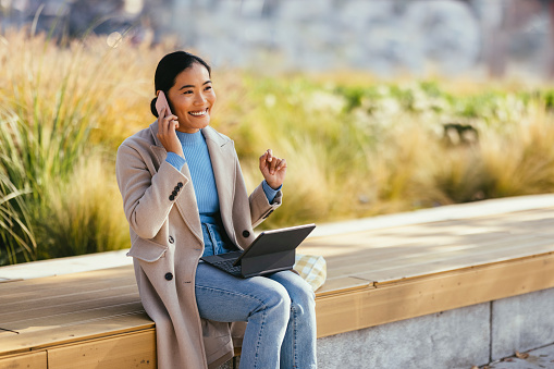 Cheerful Asian woman is sitting outside on a sunny autumn day. She has a laptop placed on her lap while she is talking on the phone. She might have decided to work outside for a bit and is having a business call or she might be taking a break at work. She might be sitting in a park.