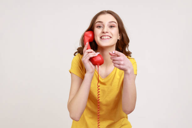Smiling attractive woman of young age in yellow t-shirt pointing finger on you holding in hands landline telephone, waiting for your call. stock photo