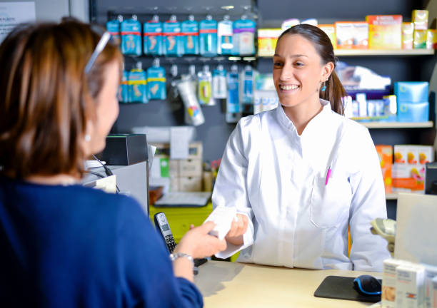 Smiling attractive young redhead pharmacist handing over prescribed medicines to an elderly female patient, view over the clients shoulder of the pharmacist stock photo
