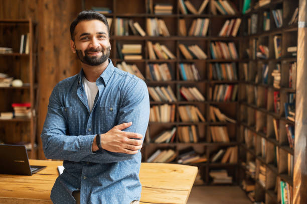 Smiling bearded indian businessman stands near desk and looks at the camera Smiling bearded indian businessman stands near desk and looks at the camera. Young positive male student in library with bookshelves on background. Proud and successful mixed-race small business owner middle eastern ethnicity stock pictures, royalty-free photos & images