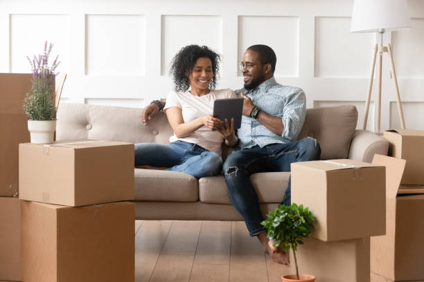 Smiling black couple use digital tablet on moving day Happy african couple husband and wife choose house removal service or search renovating ideas sit on sofa with boxes, smiling black renters owners tenants use digital tablet on moving day in new home mobile real estate stock pictures, royalty-free photos & images
