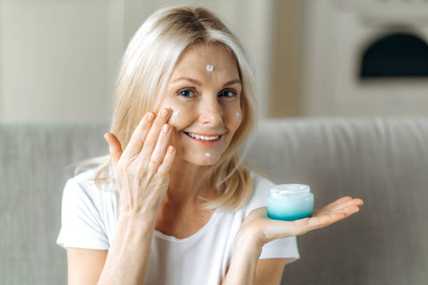 Smiling blonde senior woman apply anti-aging cream to her face. Happy mature woman using cosmetic cream to hide wrinkles on face, moisturizes and nourishes the skin stock photo
