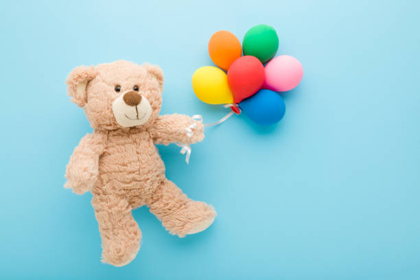 Smiling brown teddy bear holding heap of colorful balloons on light blue table background. Pastel color. Closeup. Congratulation concept. Top down view. stock photo