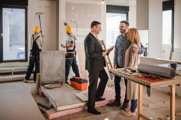 Smiling building contractor talking to a couple inside of renovating apartment. Happy mid adult investor communicating with a couple during home renovation process in the apartment. Workers are in the background. Home Equity & Renovation stock pictures, royalty-free photos & images