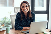 istock Smiling business woman working with laptop while looking at camera in modern startup office. 1348209421