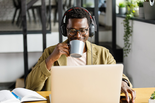 Front view of confident young businessman smiling while listening to music via headphones, sipping coffee and working at his desk at the office.
