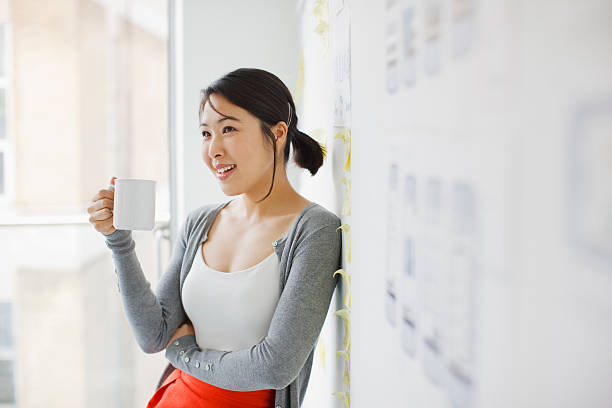smiling businesswoman leaning against whiteboard and drinking coffee - woman drinking coffee stockfoto's en -beelden