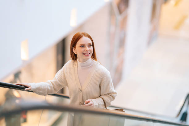 Smiling charming young woman shopaholic holding on escalator handrail and riding escalator going up in shopping mall stock photo