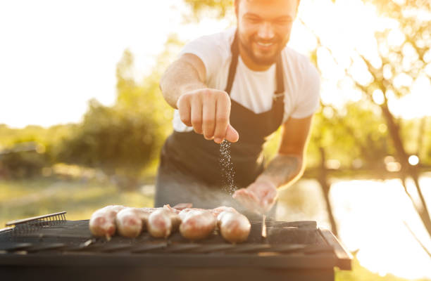 Smiling chef salting sausages on grill Crop male cook smiling and spilling salt of delicious fried sausages on grill on blurred background of countryside on sunny day plus sign photos stock pictures, royalty-free photos & images