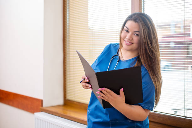 lächelt zuversichtlich ärztin in einem krankenhaus flur neben einem fenster steht, trägt eine blaue uniform und stethoskop, hält einen ordner - assistent stock-fotos und bilder