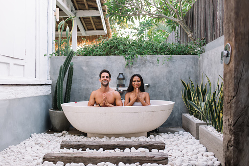 Content man and woman holding hands in namaste relaxing in outdoors bathroom while enjoying summer vacation in Bali villa