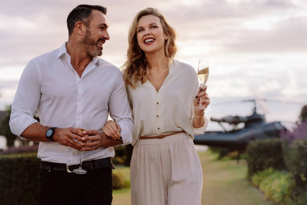 Smiling couple walking outdoors Smiling couple walking outdoors holding a glass of wine. Caucasian man and woman with a drinks walking together with a helicopter in background. high society stock pictures, royalty-free photos & images