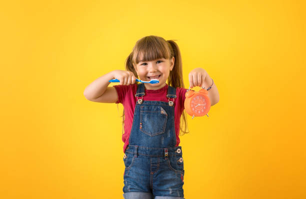 Smiling cute little girl holding alarm clock and toothbrush isolated yellow background. Brush teeth concept stock photo