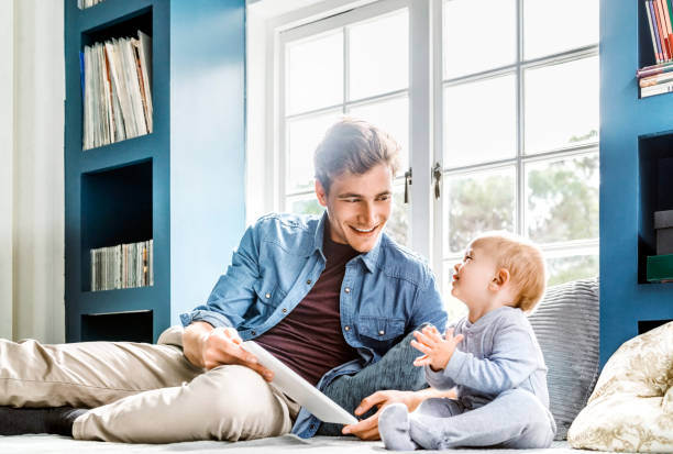 Smiling father showing digital tablet to baby Smiling father showing digital tablet to baby. Happy man sitting with toddler against window. They are at home. alcove window seat stock pictures, royalty-free photos & images