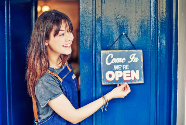 Smiling female entrepreneur hanging open sign on deli door Beautiful young entrepreneur hanging open sign on door. Smiling owner of delicatessen is standing in doorway while looking away. She is wearing apron. irish women stock pictures, royalty-free photos & images