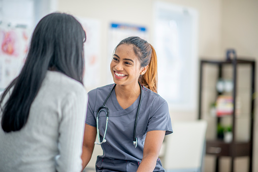 A young female nurse of Asian descent is smiling widely at her patient who is sitting across her. She is wearing medical scrubs and has a stethoscope around her neck.