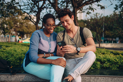 Smiling couple is sitting outdoor and looking at the phone.