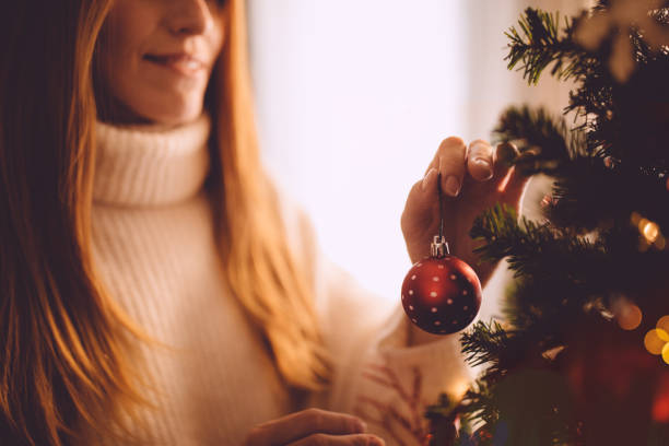 glimlachend gember vrouw versieren kerstboom met rode kerst bal - vrouw kerstboom versieren stockfoto's en -beelden