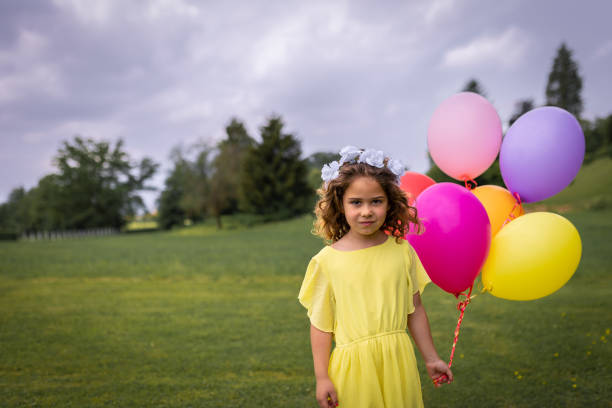 A smiling girl in spring nature in a yellow dress, with a wreath of flowers on her head and with colored balloons filled with helium. stock photo