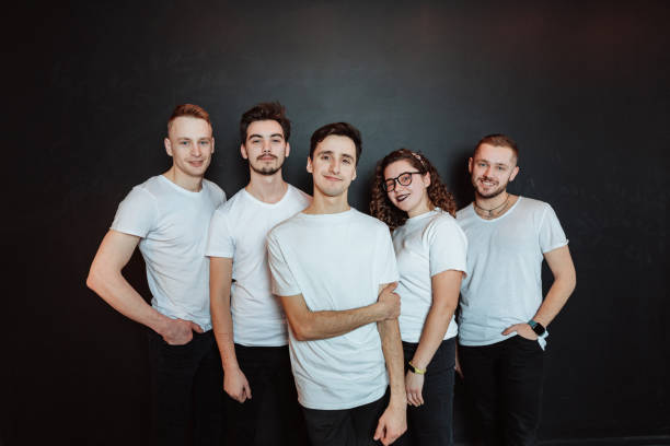 Smiling guys and girl in white t-shirts standing by black wall in front of camera Friendly smiling guys and girl in casualwear standing by black wall in front of camera and looking at you teamwork photos stock pictures, royalty-free photos & images