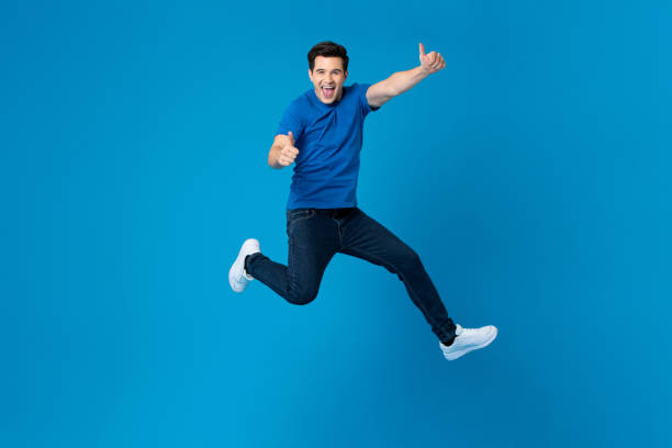 Smiling handsome American man joyfully jumping and doing double thumbs up gesture isolated on blue studio background stock photo
