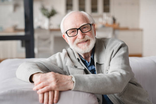 Smiling happy healthy caucasian senior old elderly man grandfather relaxing resting on the sofa couch looking at camera with toothy smile at home. Pension loan concept. stock photo