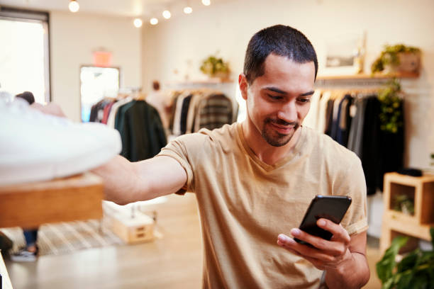 Smiling Hispanic man using smartphone in a clothes shop Smiling Hispanic man using smartphone in a clothes shop clothing store photos stock pictures, royalty-free photos & images