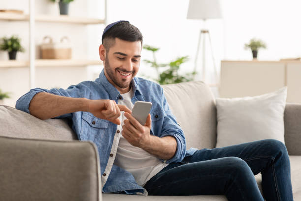 Smiling jew man using smart phone sitting on couch stock photo