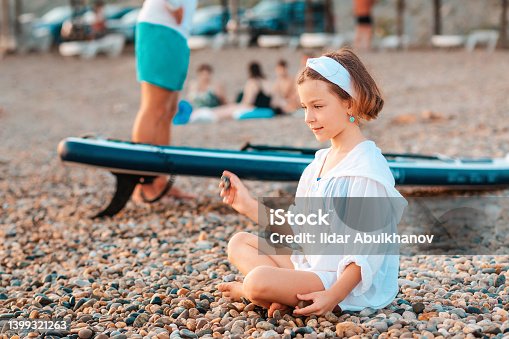 istock Smiling little girl sitting on the beach and throws a pebbles. Man with surf board on the background. Concept of summer active vacation 1399321263