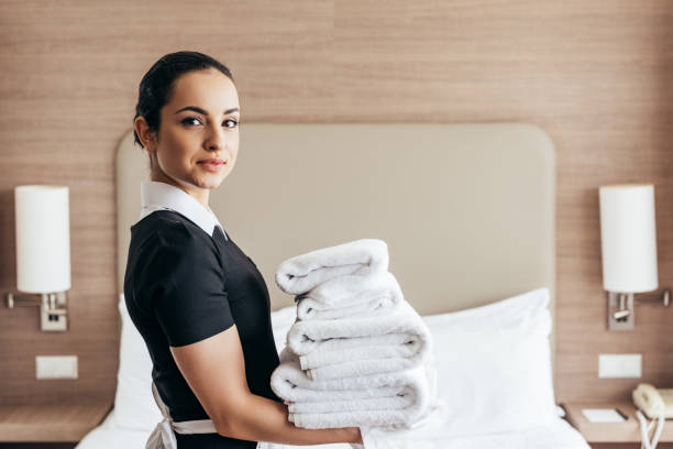 smiling maid holding pile of folded towels near bed and looking at camera in hotel room smiling maid holding pile of folded towels near bed and looking at camera in hotel room maid stock pictures, royalty-free photos & images