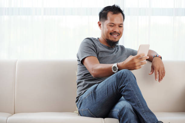 Smiling man checking his phone Filipino man sitting on sofa and reading messages in his phone filipino ethnicity stock pictures, royalty-free photos & images