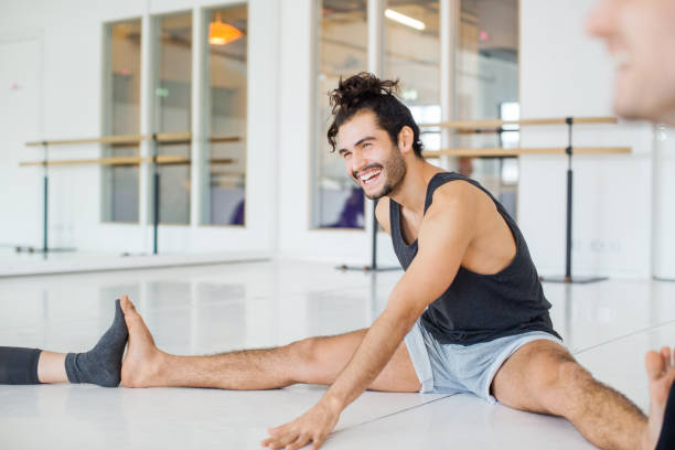 Smiling man doing stretching in ballet studio Smiling young man doing stretching exercise with friends in ballet studio. Multi-ethnic male and female dancers are warming up before practice. They are in rehearsal room. doing the splits stock pictures, royalty-free photos & images