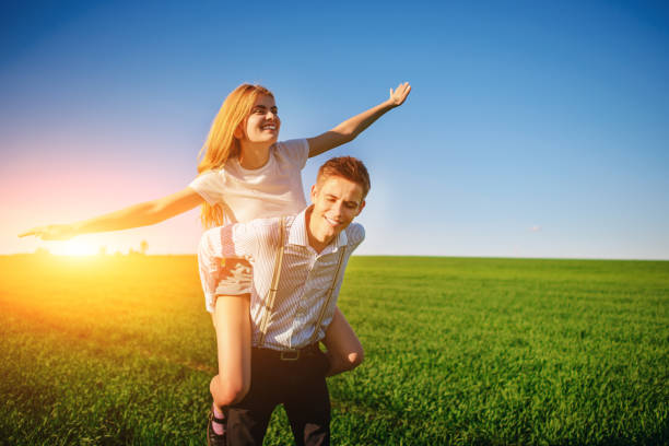 Smiling Man is holding on his back happy woman, who pulls out her arms and simulates a flight against the background of the blue sky and the green field stock photo