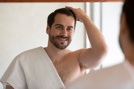Happy millennial handsome man in towel look in mirror in bathroom at home get ready in morning. Smiling young male style hair make hairdo prepare with procedures in bath. Beauty concept.