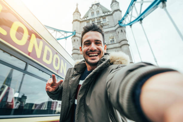 Smiling man taking selfie portrait during travel in London, England - Young tourist male taking memory pic with iconic england landmark - Happy people wandering around Europe concept stock photo