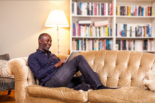 Mature man portrait in living room, smiling and reading on a digital tablet computer, sitting on a couch. Typical cosy english living room with library in the background. Positive scene with warm colors.