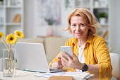 Smiling mature blond businesswoman with smartphone looking at you while working remotely in front of laptop on quarantine