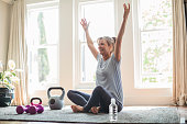 Smiling mature woman with arms raised doing yoga. Fit female is sitting by exercise equipment on rug. She is wearing sports clothing at home.