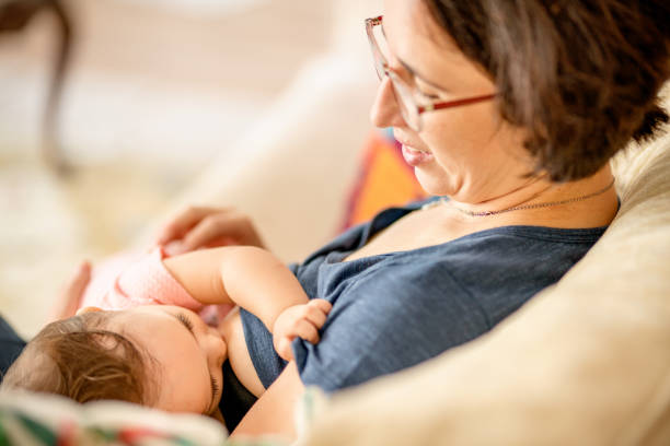 Smiling mom breastfeeding her little girl on a sofa at home Smiling mother breastfeeding her little daughter while sitting on a living room sofa at home increase breast milk stock pictures, royalty-free photos & images