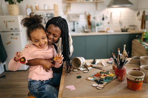 Smiling mid adult mother with dreadlocks embracing daughter holding craft products by table at home